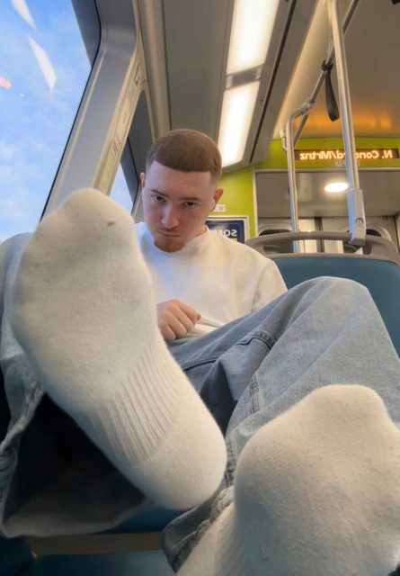 A young man with a buzzcut sitting on public transit extends his white socked feet directly toward the camera lens in a low-angle POV shot.