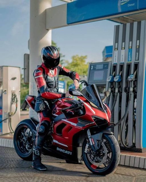A rider clad in a full red and black leather suit and helmet sits on a red Ducati motorcycle at a gas station.