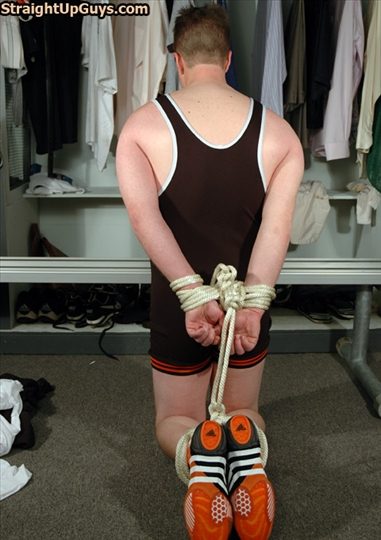 A man wearing a wrestling singlet kneels in a locker room with his wrists and ankles bound together behind his back with rope.