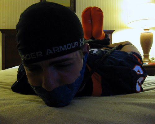 A young man in a football jersey and orange socks lies bound and gagged with blue duct tape on a bed.