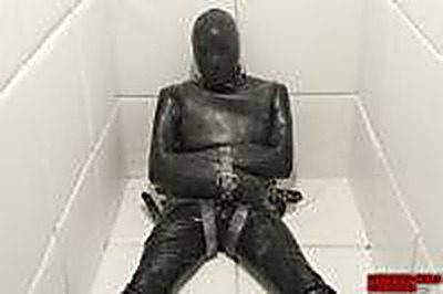 A person wearing a full black latex suit and hood is bound in straps inside a white tiled room.