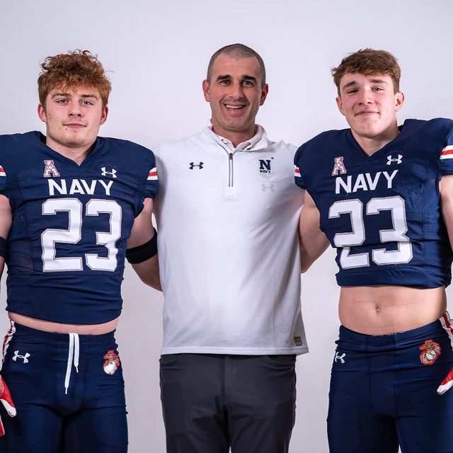 A mature coach stands with his arms around two young football players wearing padded Navy jerseys and tight athletic pants.