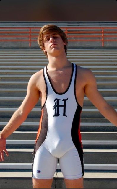A young, athletic man poses in a tight white and black wrestling singlet in front of stadium bleachers.