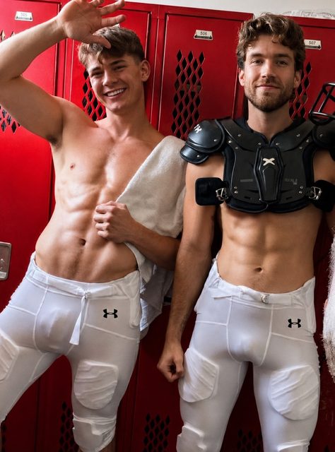 Two fit young men in tight padded white football pants pose together in a red locker room, with one wearing black shoulder pads.