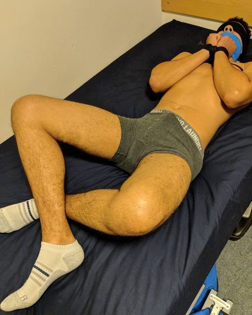 A blindfolded and gagged man with his wrists bound lies on a blue bed wearing grey boxer briefs and socks.