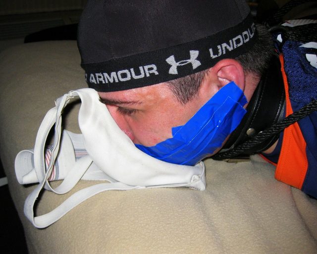 A bound man wearing a leather collar and tape gag lies face down with his nose in a white jockstrap.