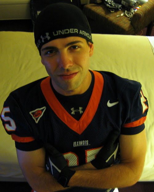 A young man posing in a full Illinois football jersey, gloves, and compression skull cap in a bedroom setting.