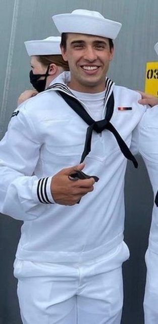 A fit young man smiles while wearing a traditional white US Navy sailor uniform and hat.