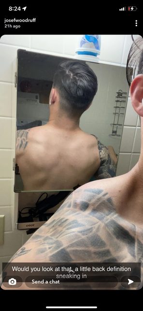 A muscular man with extensive tattoos on his shoulders and arm takes a mirror selfie showing his back definition in a bathroom.