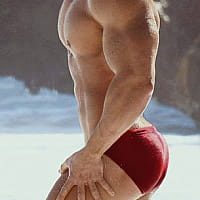 A side profile view of a muscular, smooth-skinned man posing outdoors in red briefs.