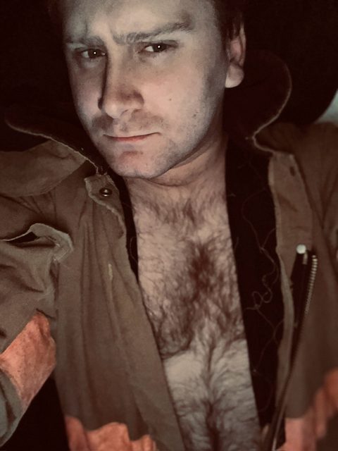A man with a dense mat of chest hair poses in an unzipped tan flight suit.