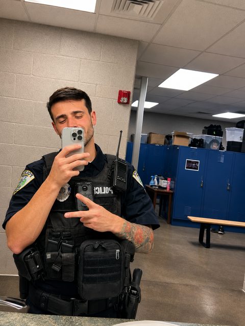 A tattooed man in a full tactical police uniform takes a mirror selfie inside a locker room.