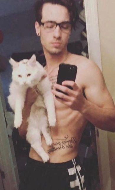A shirtless young man with glasses and abdominal tattoos takes a mirror selfie while holding a large white cat.