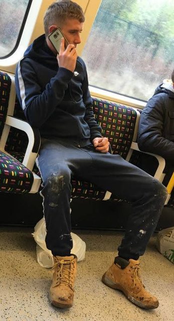 A young man in a paint-stained navy tracksuit and tan work boots sits on a train while talking on his phone.