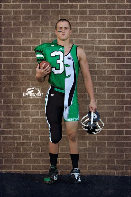 A muscular man poses against a brick wall in a split football and wrestling uniform.