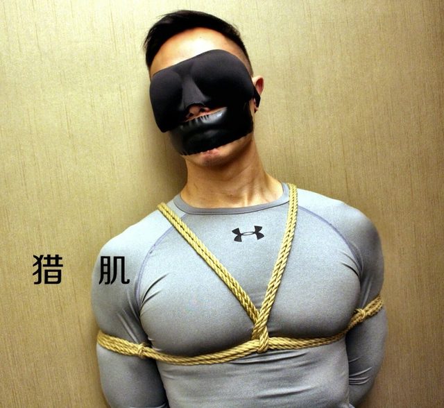 A muscular Asian man wearing a grey compression shirt is tied with rope bondage, blindfolded, and gagged against a wall.