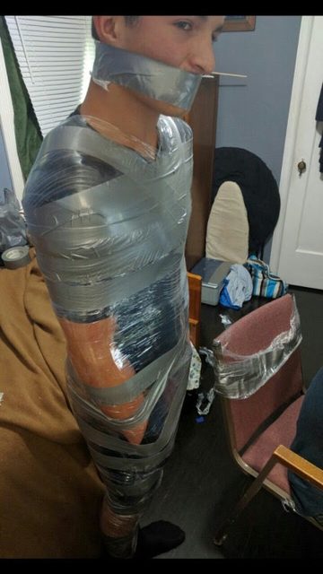 A man stands bound in a domestic setting, his entire body and mouth wrapped in layers of clear plastic film and silver duct tape.