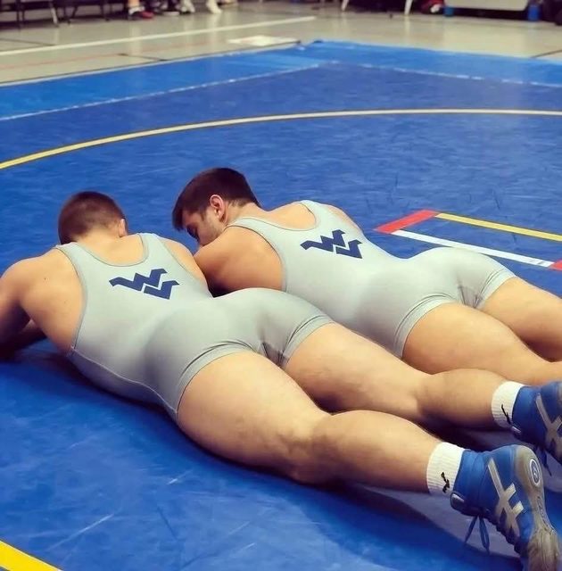 Two muscular men in grey wrestling singlets lie face down on a blue wrestling mat.
