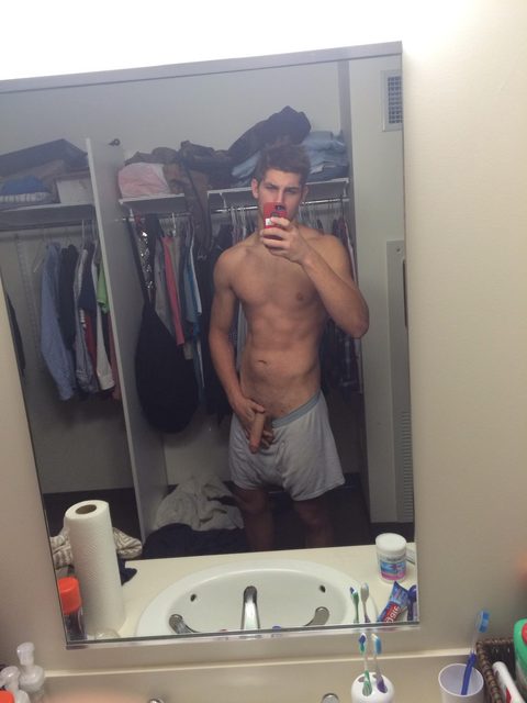 A shirtless young man takes a mirror selfie in a bathroom with his underwear pulled down to reveal his penis.