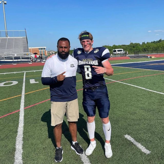 A bearded man in casual athletic wear poses next to a young football player in a full navy uniform on a sunlit field.