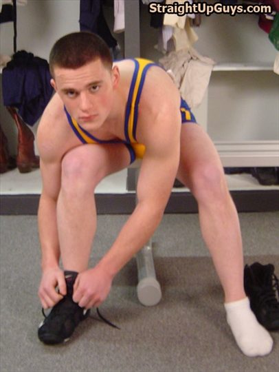 A young man with a buzzcut wears a blue and yellow wrestling singlet while tying his shoes in a locker room.