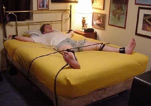 A man is restrained in a spread-eagle position on a bed using leather cuffs and rope attached to the bed frame.