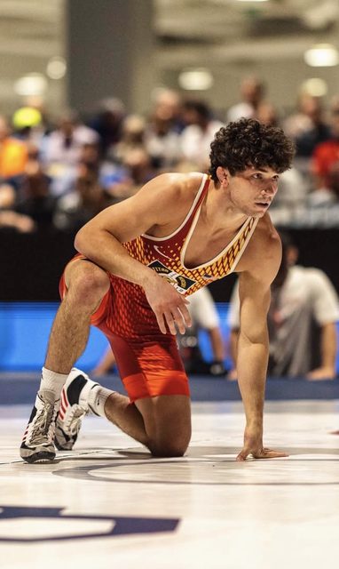 A muscular young jock in a red patterned wrestling singlet kneels on a wrestling mat in an arena.