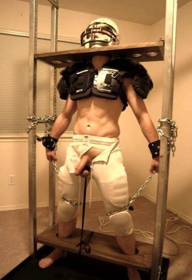 A toned man wearing football shoulder pads and a helmet stands locked in a tall wooden pillory frame with exposed genitals.