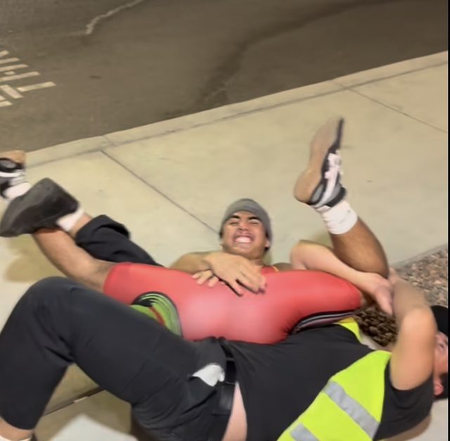 Two young men engage in an amateur wrestling match on a sidewalk at night, with one wearing a red wrestling singlet.