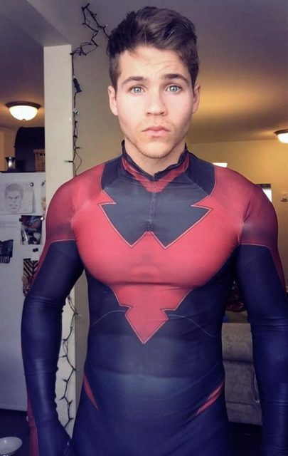 A young muscular man poses in a tight red and dark blue full-body suit indoors.