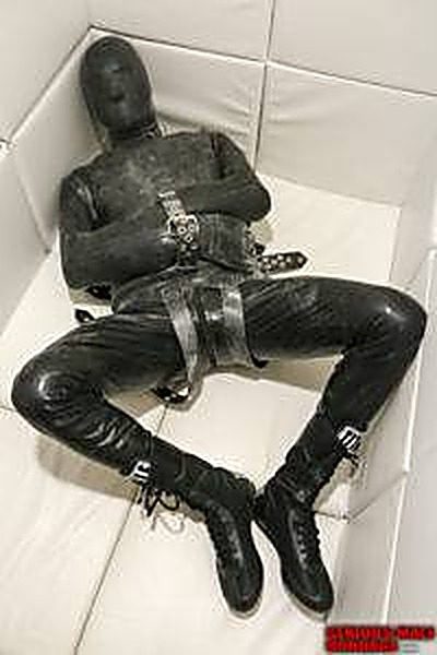 A subject fully encased in a black latex gimp suit and boots lies bound in heavy straps within a white padded cell.