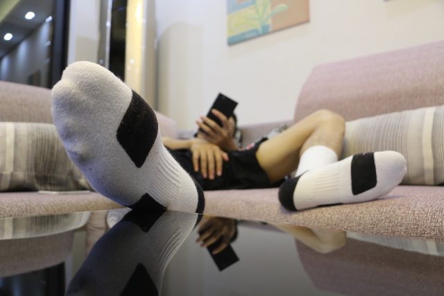 A person wearing white athletic socks and black gym shorts relaxes on a sofa while looking at a phone.