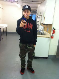 A young man in a black Nike hoodie and camouflage pants takes a mirror selfie in a utility room setting.