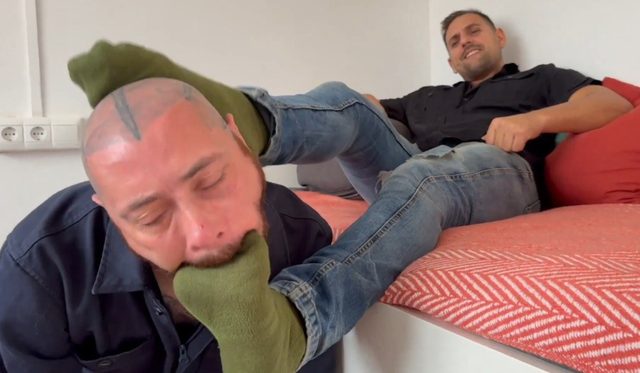 A bearded man with a tattooed head kneels to suck on the green socks of a man reclining on a bed in a show of submissive worship.