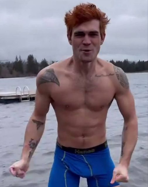 A muscular red-headed man with tattoos stands shirtless in a lake wearing blue compression shorts.