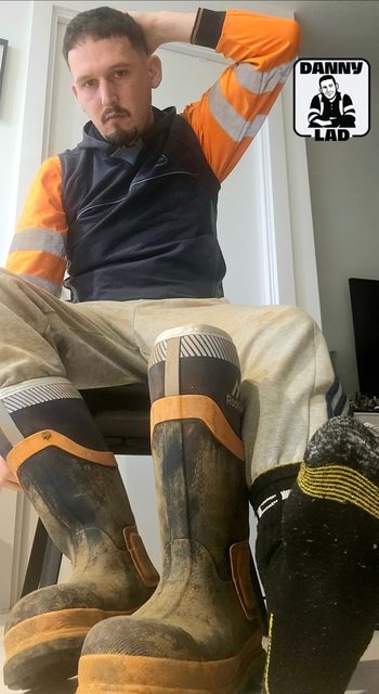 A bearded man in a high-visibility construction uniform sits with his mud-stained rubber boots and a socked foot prominently displayed.