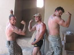 Three shirtless men in military-style camo pants and shorts flex their muscles in a group pose.