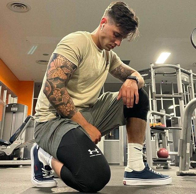 A muscular man with arm tattoos kneels in a gym wearing compression leggings under athletic shorts and white crew socks.