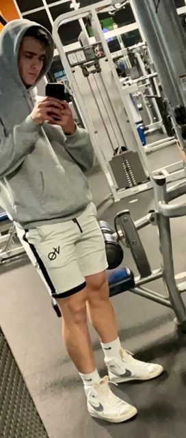 A young man in a grey hoodie and short gym shorts takes a mirror selfie inside a gym.