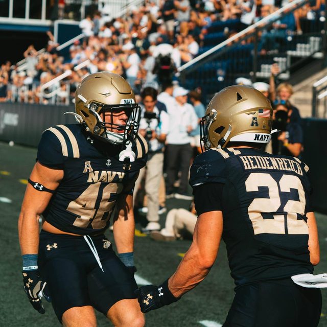 Two muscular men in full Navy football kits with gold helmets face each other on an outdoor field.