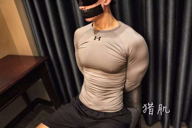 An athletic man wearing a tight compression shirt and gym shorts sits blindfolded and mouth-taped with his hands bound behind his back in a hotel room.