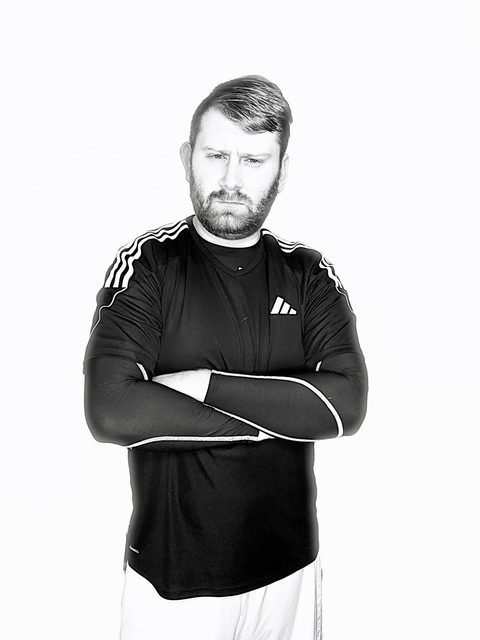 A bearded man wears a black Adidas jersey and compression sleeves with his arms crossed against a plain white background.