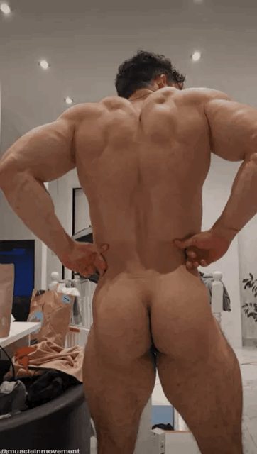 A muscular bodybuilder stands naked and flexes his back and glutes for the camera in an indoor setting.