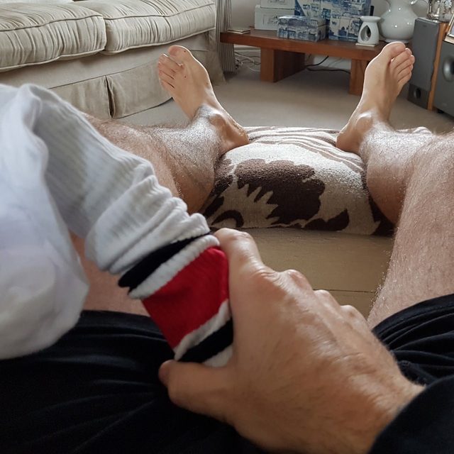 A point-of-view shot shows a hairy-legged man in black gym shorts holding a striped tube sock over his lap while sitting in a living room.