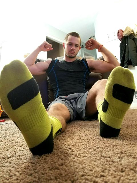 A muscular man in athletic wear sits on the carpet with his socked feet extended toward the camera while flexing his biceps.