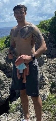 A shirtless tattooed young man in grey gym shorts stands on a rocky coastline.
