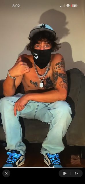 A shirtless, tattooed young man wearing a face mask and light blue jeans sits on a couch.