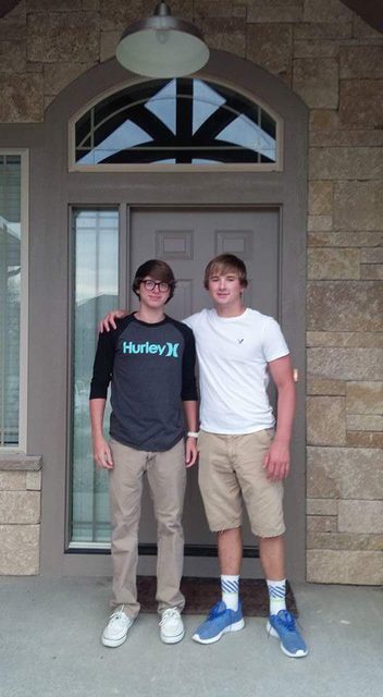 Two young men stand together in front of a house.
