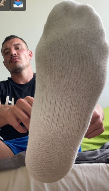 A man with a buzzcut and mustache presents the sole of his white cotton sock to the camera while sitting on a bed.