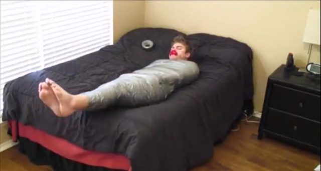 A young man lies on a bedroom bed, tightly cocooned in silver duct tape from shoulders to feet with a red gag in his mouth.